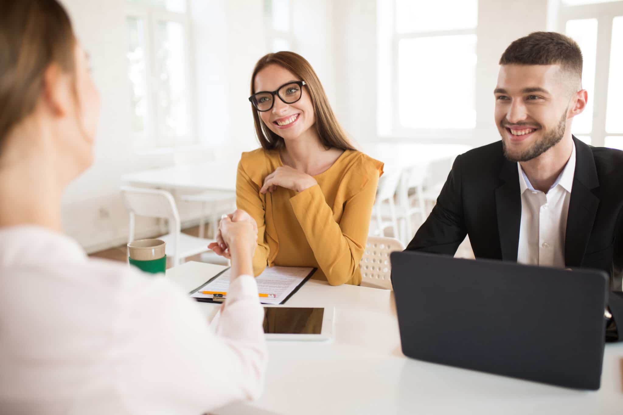 Smiling business woman in eyeglasses happily shaking applicant hand with business man near. Young joyful employers spending job interview in modern office